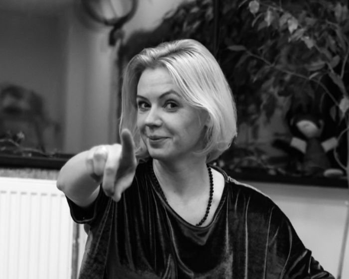 Black-and-white portrait of a blonde woman pointing at the camera with a playful smile, indoors.