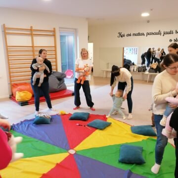 Parents and children in a bright playroom gathered around a colorful parachute mat for a group activity.