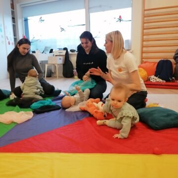 Adults and young children on a colorful play mat in a bright room, interacting and playing with babies.