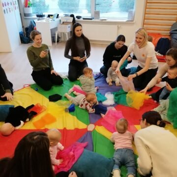 Mothers and babies sit on a large rainbow mat, playing together in a bright playroom.