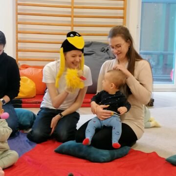 Adults and a child sit on a colorful mat in a playroom; one person wears a penguin hat and holds a yellow plush chick puppet.