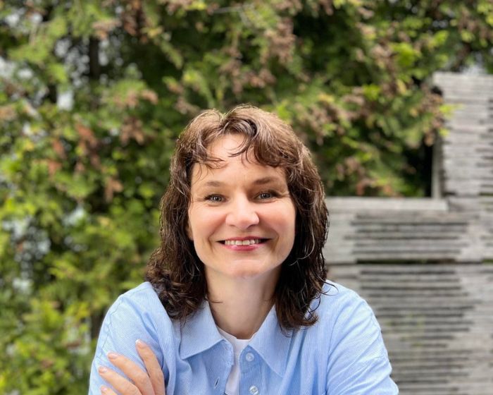Smiling woman with shoulder-length brown hair outdoors, wearing a light blue shirt, arms crossed in front of greenery and steps.