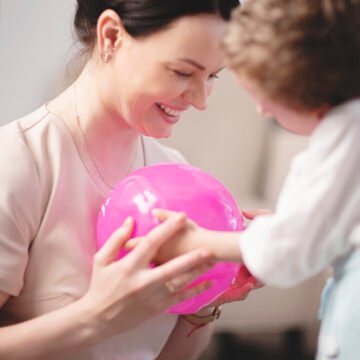 Woman smiling while holding a pink balloon, playing with a child indoors.