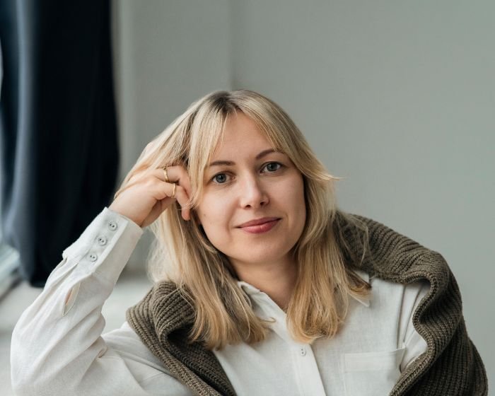 Portrait of a woman with shoulder-length blonde hair wearing a white blouse and a brown cardigan draped over her shoulders, smiling softly.