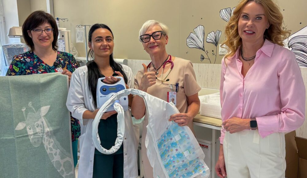 Four healthcare workers in a hospital room pose with a newborn in a bassinet, smiling at the camera.