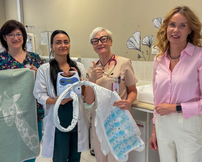 Four healthcare workers in a hospital room pose with a newborn in a bassinet, smiling at the camera.