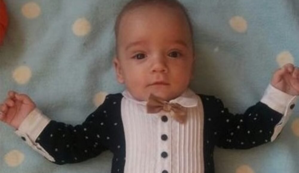 Infant lying on a blue polka-dot blanket, wearing a white pleated shirt, navy cardigan with white dots, and a beige bow tie.