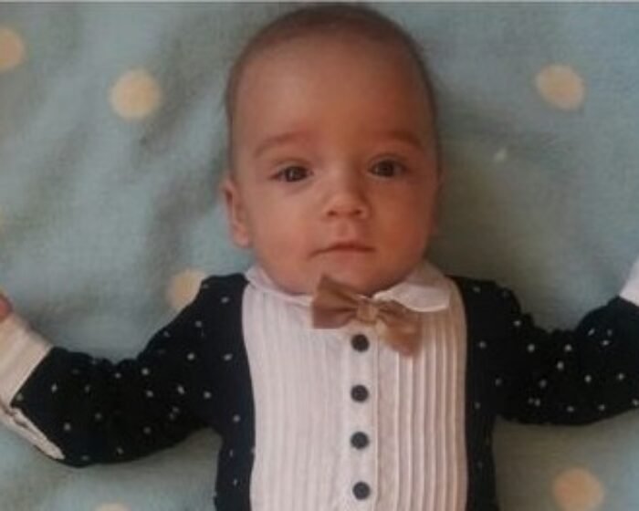 Infant lying on a blue polka-dot blanket, wearing a white pleated shirt, navy cardigan with white dots, and a beige bow tie.