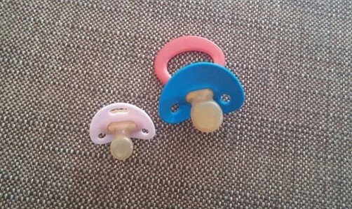 Two baby pacifiers with beige nipples, one pink ring and one blue ring, resting on a gray woven fabric surface.