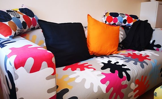 Colorful sofa with abstract splash-pattern upholstery, black and orange throw pillows, and a dark garment draped on the right.