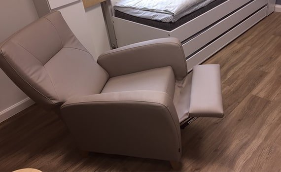 Beige reclining armchair with extended footrest in a bedroom with wood flooring and a bed in the background.