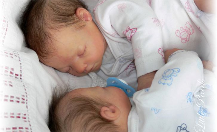 Two newborn babies sleeping close together, one with a blue pacifier resting beside the other on a white blanket.