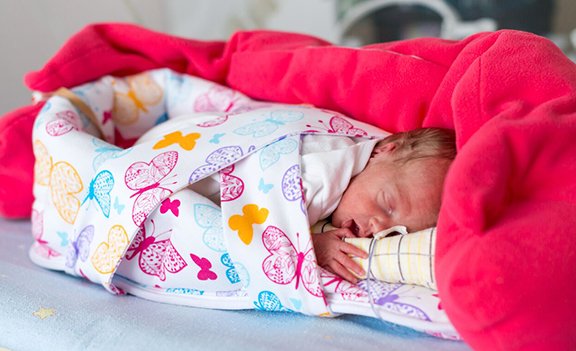Newborn baby sleeping wrapped in a colorful butterfly-pattern blanket under a pink coverlet on a bed.