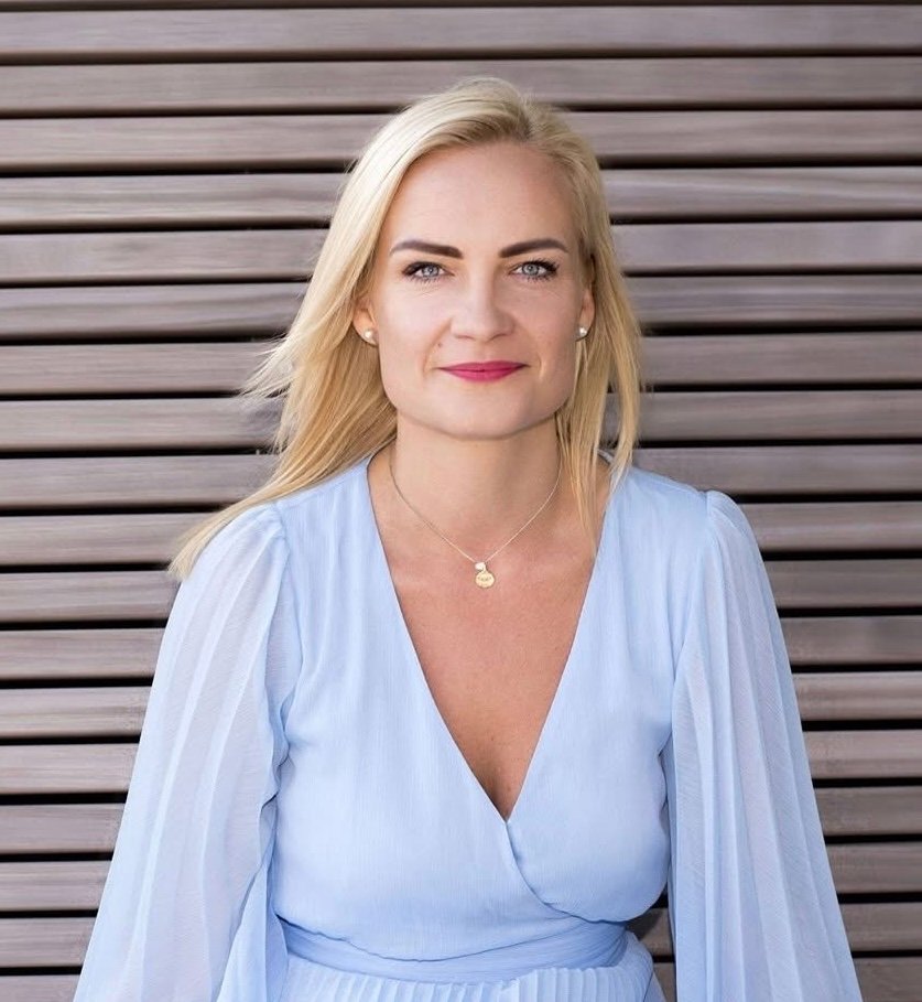 Portrait of a blonde woman in a light blue dress with a pendant necklace, smiling at the camera against a wooden slat background.