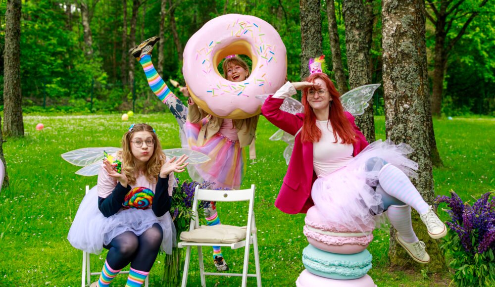 People in whimsical costumes pose outdoors with a giant pink donut prop and colorful outfits in a grassy park.