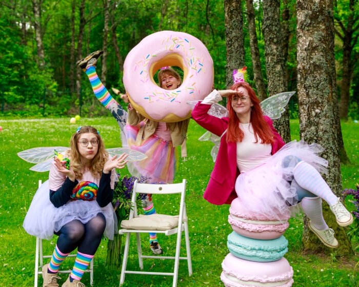 People in whimsical costumes pose outdoors with a giant pink donut prop and colorful outfits in a grassy park.