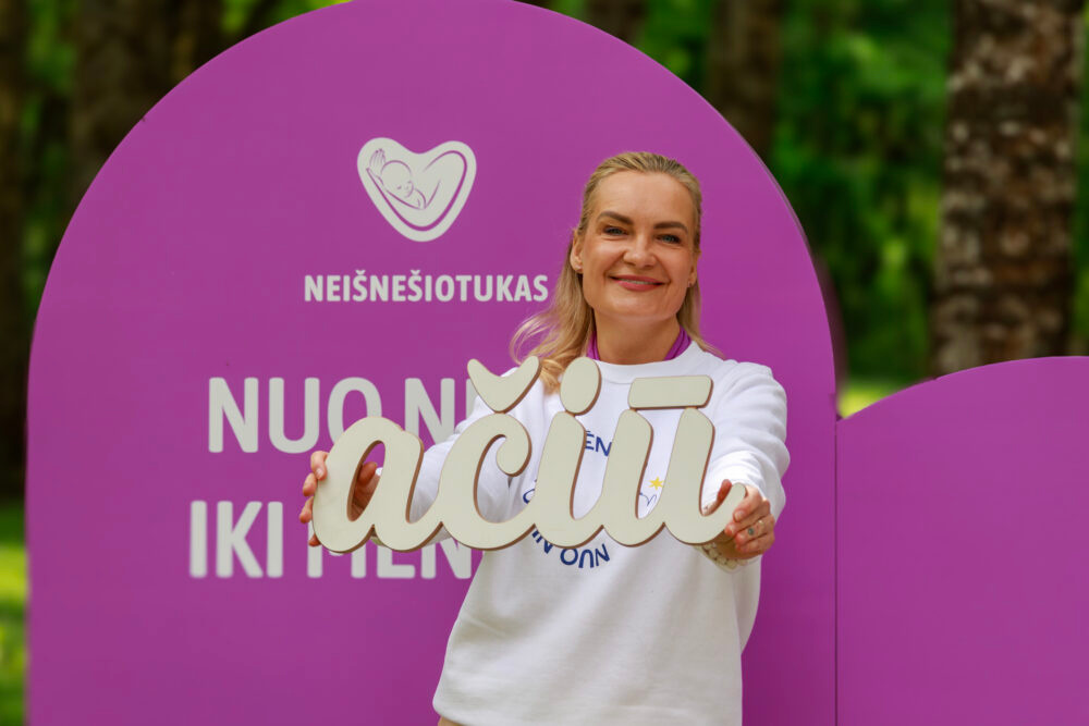 Smiling woman in a white shirt holds a large decorative sign in front of a purple circular backdrop with a baby logo and text behind her.