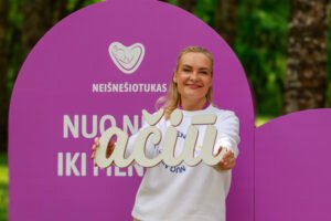 Šypsanti moteris, vilkinti baltais marškiniais, rankose laiko didelę dekoratyvinę lentelę. Už jos matomas violetinis apvalus fonas su kūdikio logotipu ir užrašu.