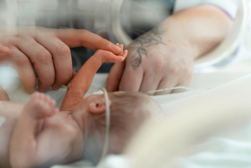 Close-up of a newborn baby's tiny fingers holding an adult's finger in a hospital setting with an IV line nearby.