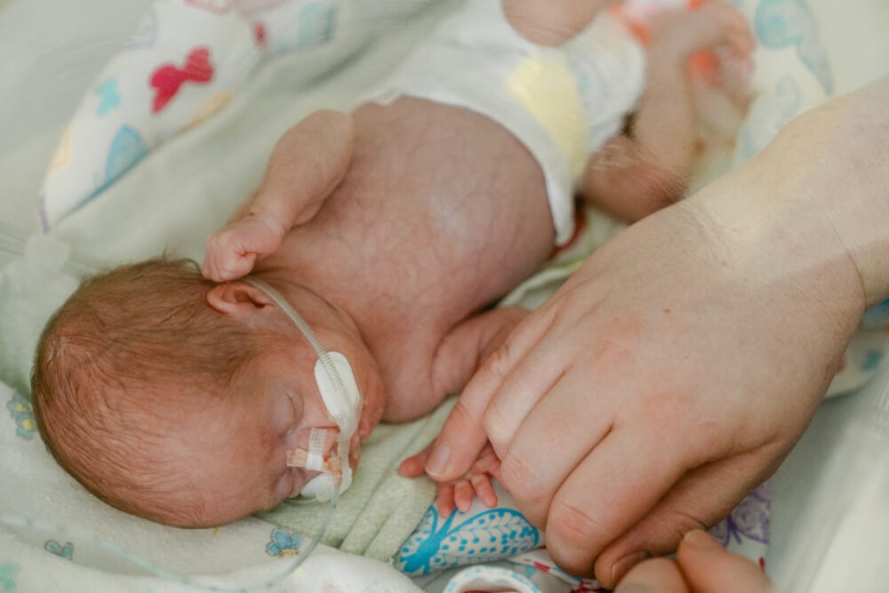 Close-up of a newborn baby resting, small hand near a parent's hand, with hospital tubing visible and a patterned blanket in the background.