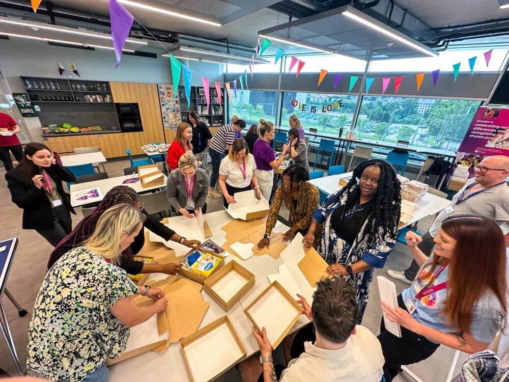 Group of people around tables in a bright classroom, assembling cardboard boxes and craft materials for a workshop under colorful bunting.