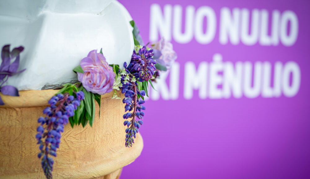 Close-up of a white statue with a purple flower wreath draped around it, against a purple background with blurred white text.