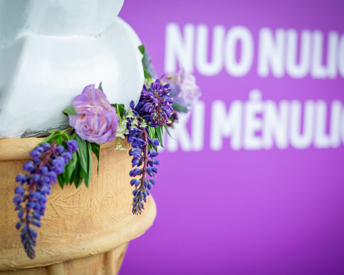 Close-up of a white statue with a purple flower wreath draped around it, against a purple background with blurred white text.
