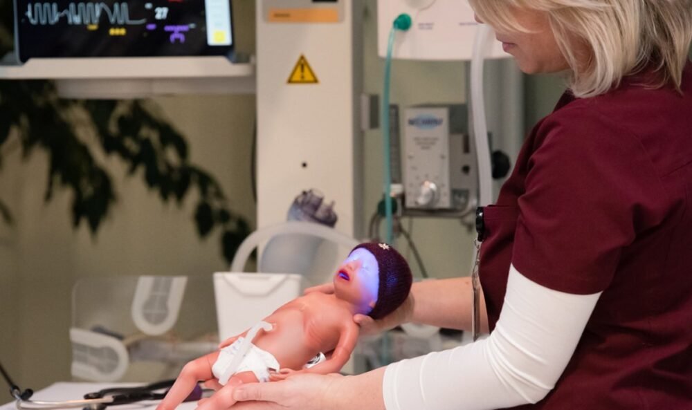 Healthcare worker in maroon scrubs holding a newborn mannequin during medical training in a hospital setting with monitors nearby.