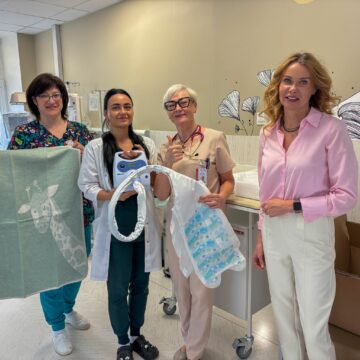 Four female healthcare workers pose for a photo in a hospital room, with medical equipment and a patient bed nearby, all smiling.