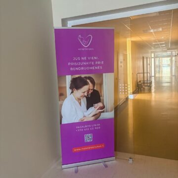 Purple promotional banner in a hospital hallway, featuring a white baby logo and Lithuanian text.