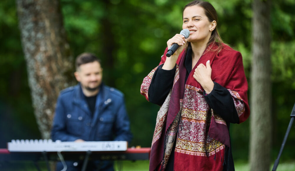 Woman singing into a microphone outdoors with a keyboard player in the blurred background.