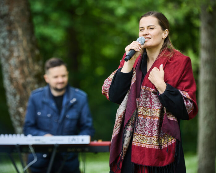 Woman singing into a microphone outdoors with a keyboard player in the blurred background.