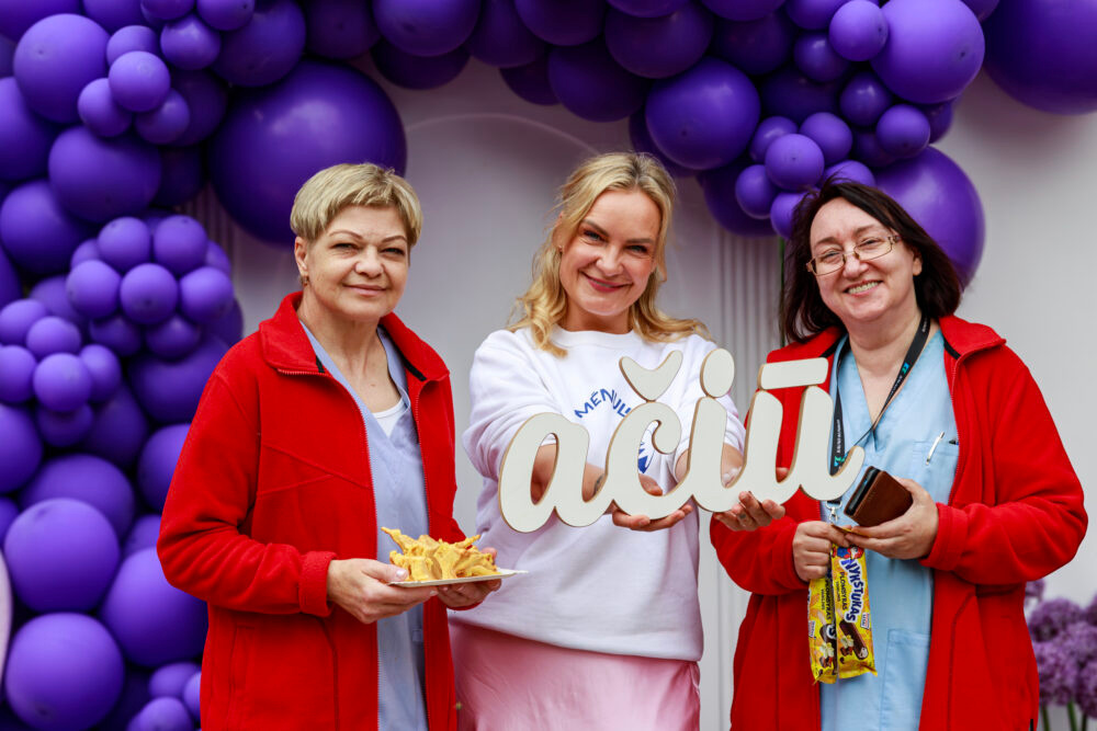 Three women in red jackets smile at the camera, posing with a large decorative cutout against a purple balloon arch backdrop (one holds chips).