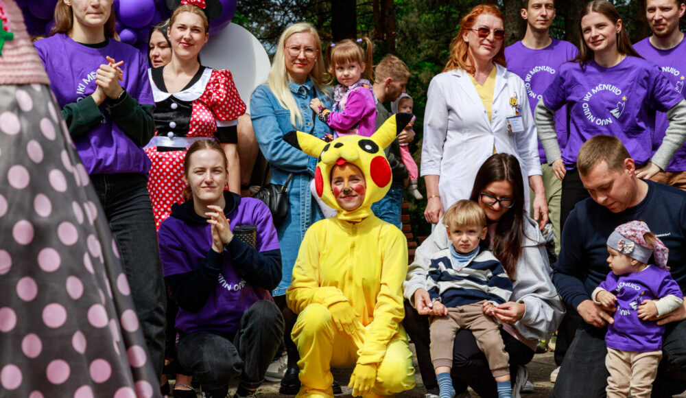 Outdoor group photo at a festival: people in purple shirts, a child in a Pikachu costume center, others smiling to the sides.