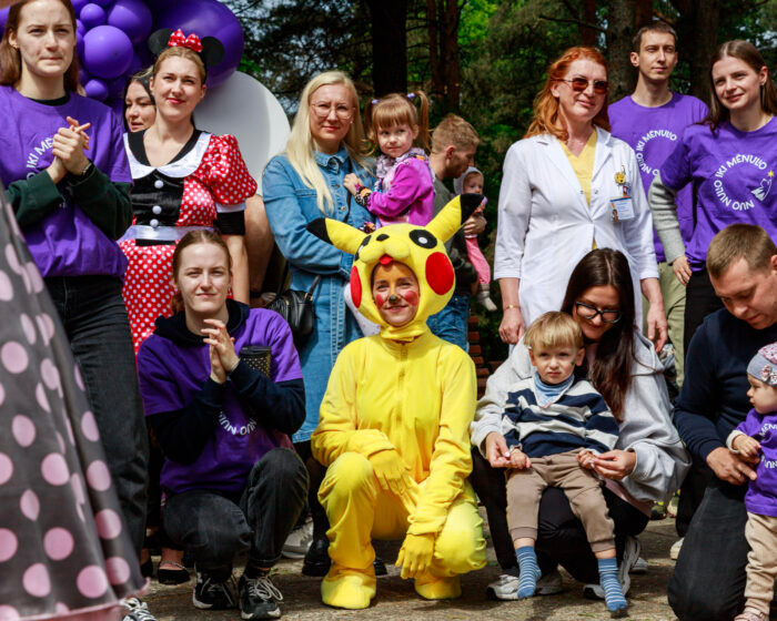 Outdoor group photo at a festival: people in purple shirts, a child in a Pikachu costume center, others smiling to the sides.