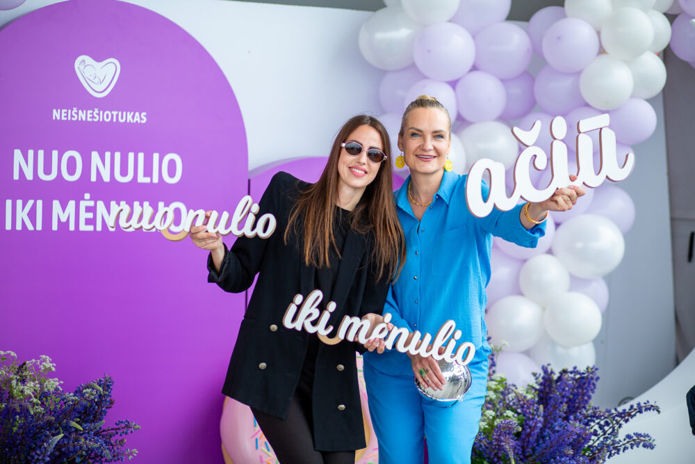 Two smiling women pose together at a celebration with a purple backdrop and lavender balloons, holding large white script cutouts.