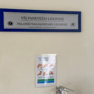 Blue bulletin board holding a tall hand-hygiene instruction poster with illustrations of hands and steps, on a beige wall across from a metal handrail attachment.