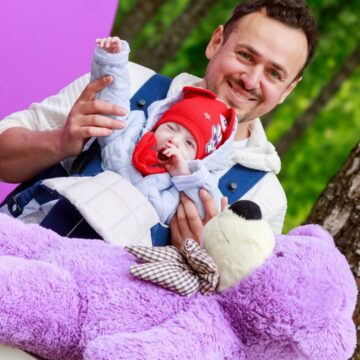 Man smiles while holding a baby in a front carrier outdoors; a large purple teddy bear rests nearby.