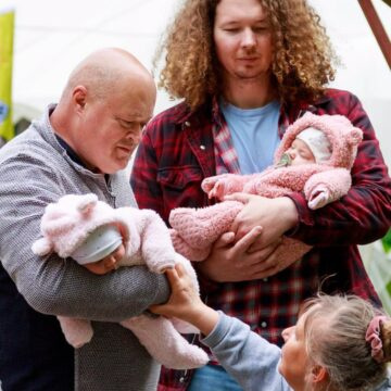 A man with a baby wearing a pink plush suit and a woman reaching to touch the baby, with another man holding a second pink-clad infant nearby.