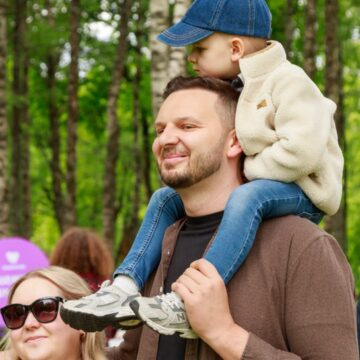 Man carrying a child on his shoulders in a wooded park; both smiling.