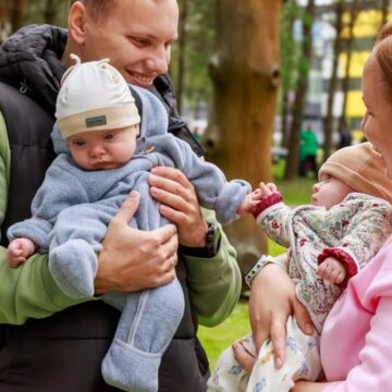 Father holds a baby in a blue onesie while a mother shows a baby toward him in a park.