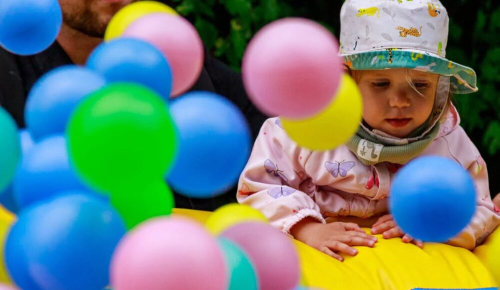 Father and toddler surrounded by colorful balloons outdoors in a play area.