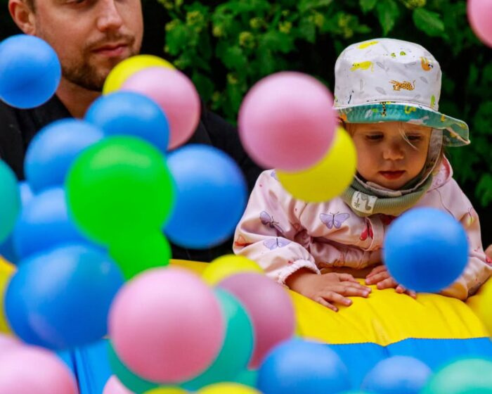 Father and toddler surrounded by colorful balloons outdoors in a play area.