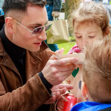 Man in sunglasses and a brown jacket feeds a child from a paper cup at an outdoor park event.