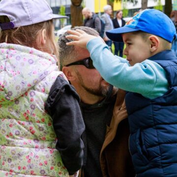 Father with sunglasses kneels outdoors as two children touch his head in a playful moment in a park.