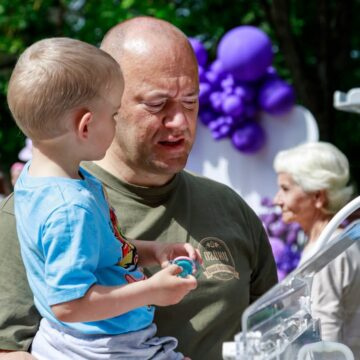 Father holding a young boy outdoors at a celebration, with purple balloons in the background.