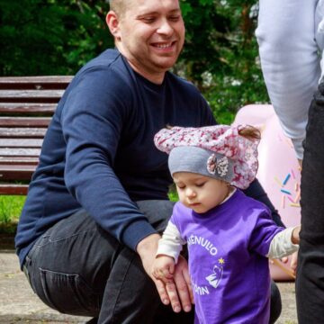 Man kneels beside a toddler girl, guiding her as she takes a step outdoors; a pink inflatable toy is in the background, and both look cheerful.