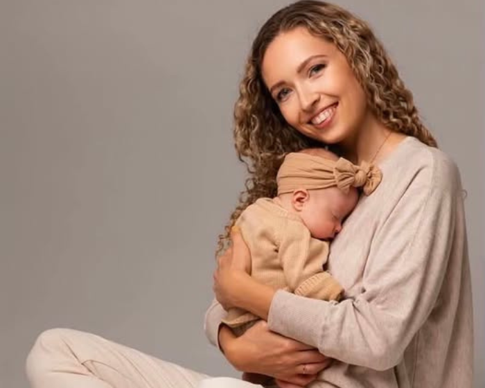 Mama švelniai glaudžia miegantį kūdikį ant rankų ir šypsosi fotoaparato link studijinėje aplinkoje.