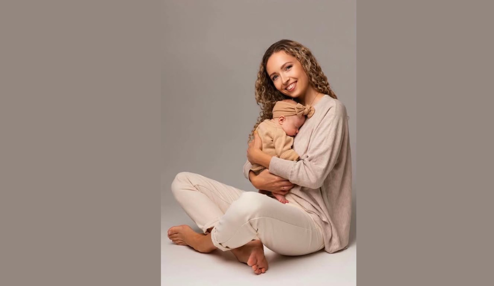 Mama švelniai glaudžia miegantį kūdikį ant rankų ir šypsosi fotoaparato link studijinėje aplinkoje.