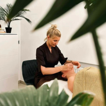 Esthetician giving a facial to a client lying on a treatment bed in a bright spa room with plants nearby.